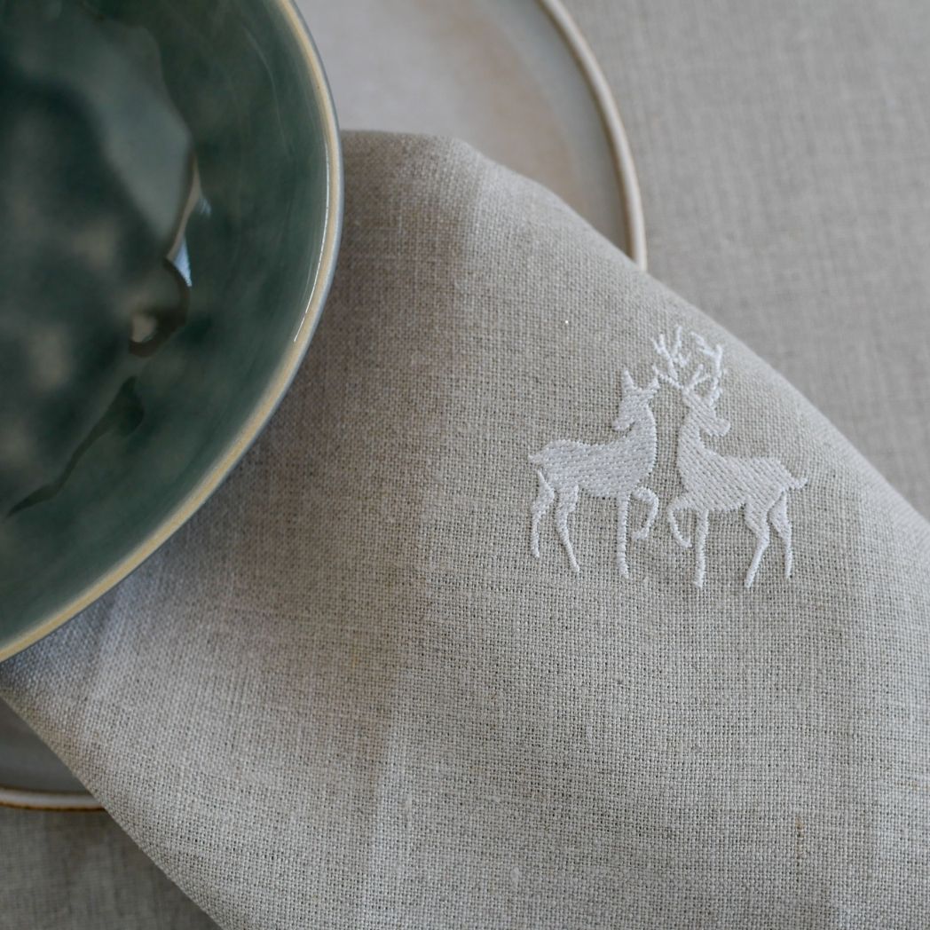 Suchen Sie schöne Leinenservietten? Auf diesem Foto handelt es sich um eine beige Leinenserviette mit einem entzückenden, weiß gestickten Hirschmotiv. Perfekt für einen Wintertisch mit rustikalem Charme!