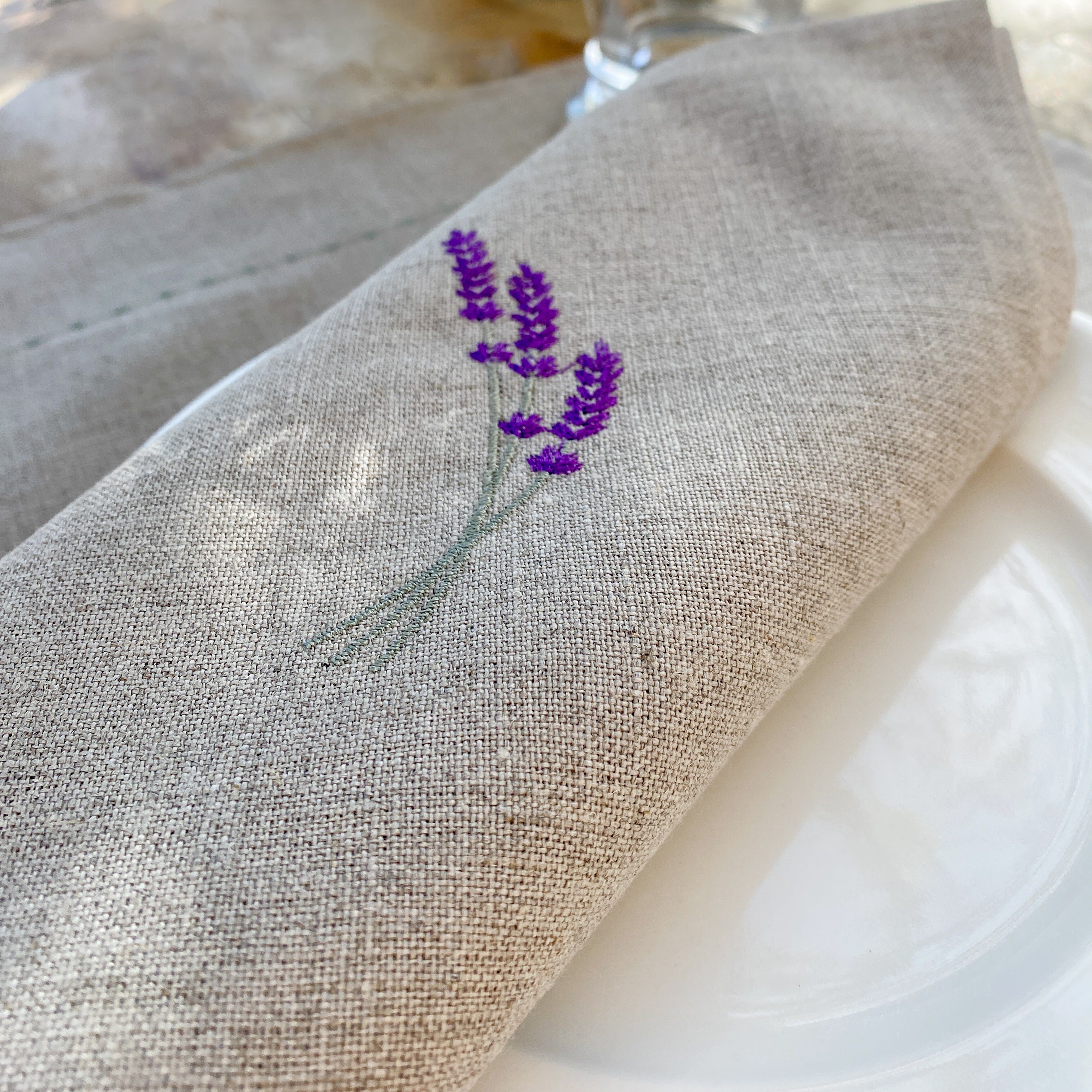 Leinenserviette mit zarter Lavendelstickerei, handgefertigt in unseren Werkstätten in Südfrankreich. Präsentiert auf einem stilvoll gedeckten Tisch – ideal für eine natürliche, elegante Tischdekoration.