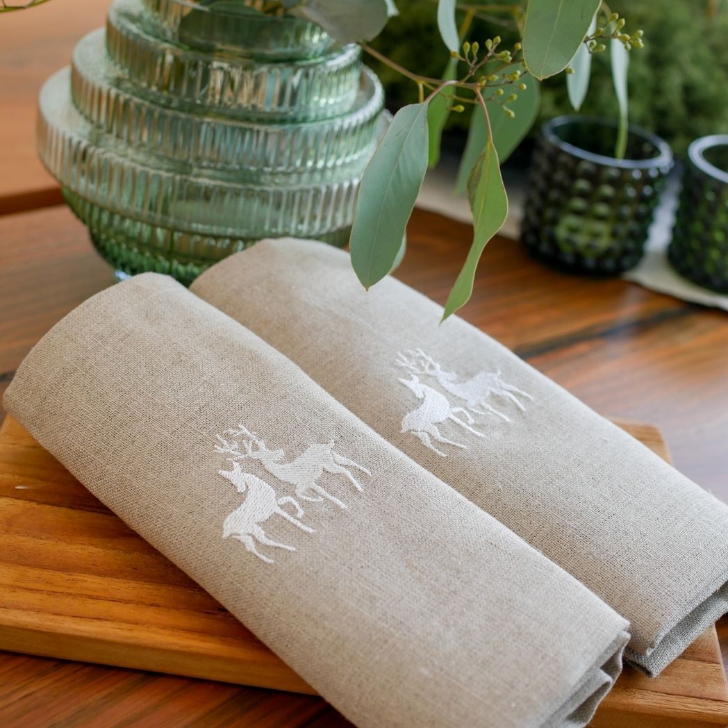 Suchen Sie schöne Leinenservietten? Auf diesem Foto handelt es sich um eine beige Leinenserviette mit einem entzückenden, weiß gestickten Hirschmotiv. Perfekt für einen Wintertisch mit rustikalem Charme!
